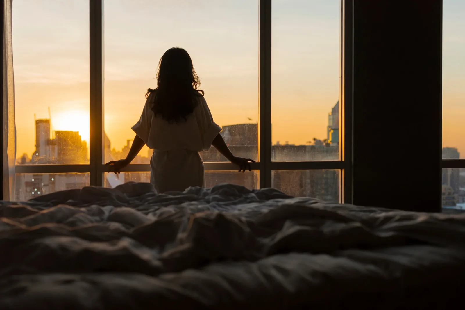 Eine Frau steht als Silhouette an einem Panoramafenster, dahinter geht die Sonne über einer Stadt auf.