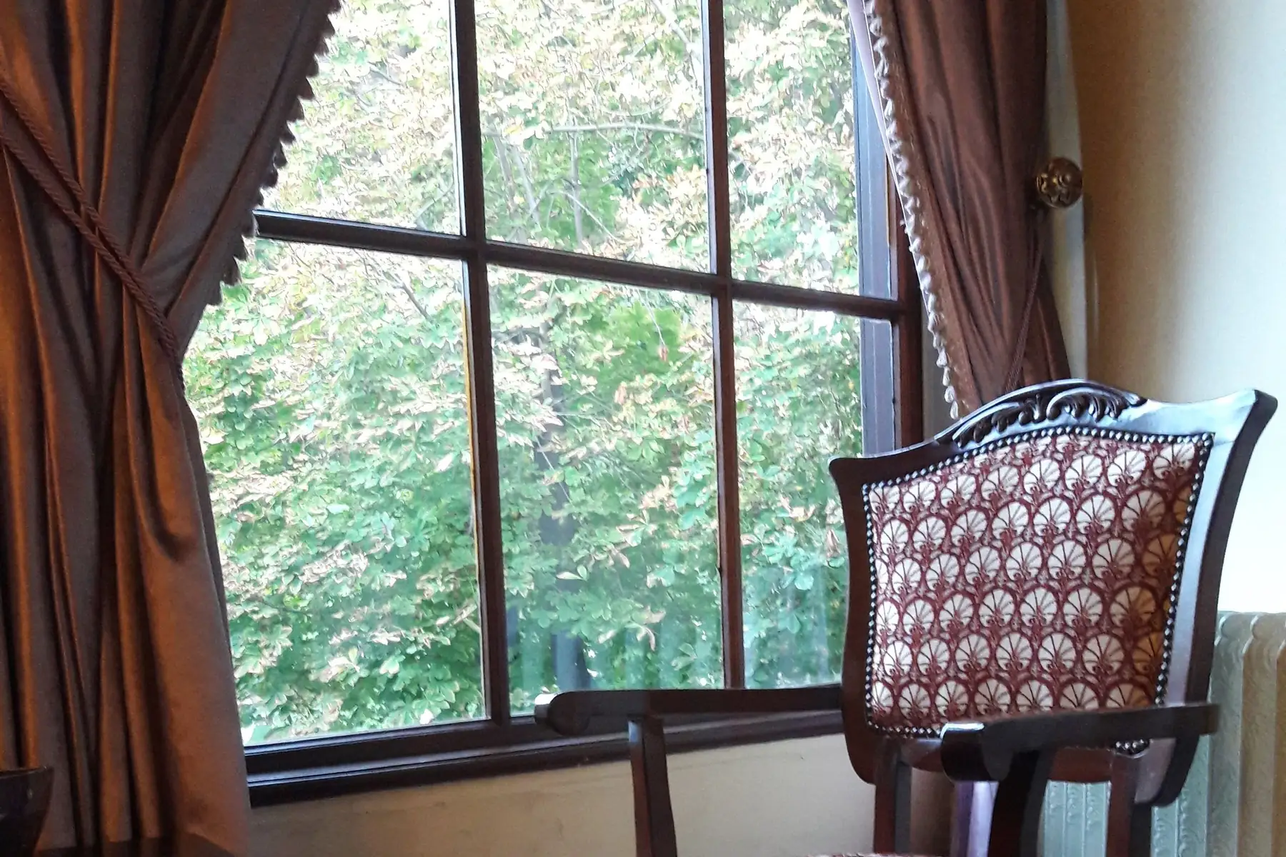 Ein gepolsterter Stuhl vor einem großen Fenster, davor ein schwerer Vorhang.